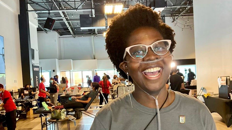 Black woman with white glasses and wearing a nasal cannula smiles in the foreground of a photo with many people in the background