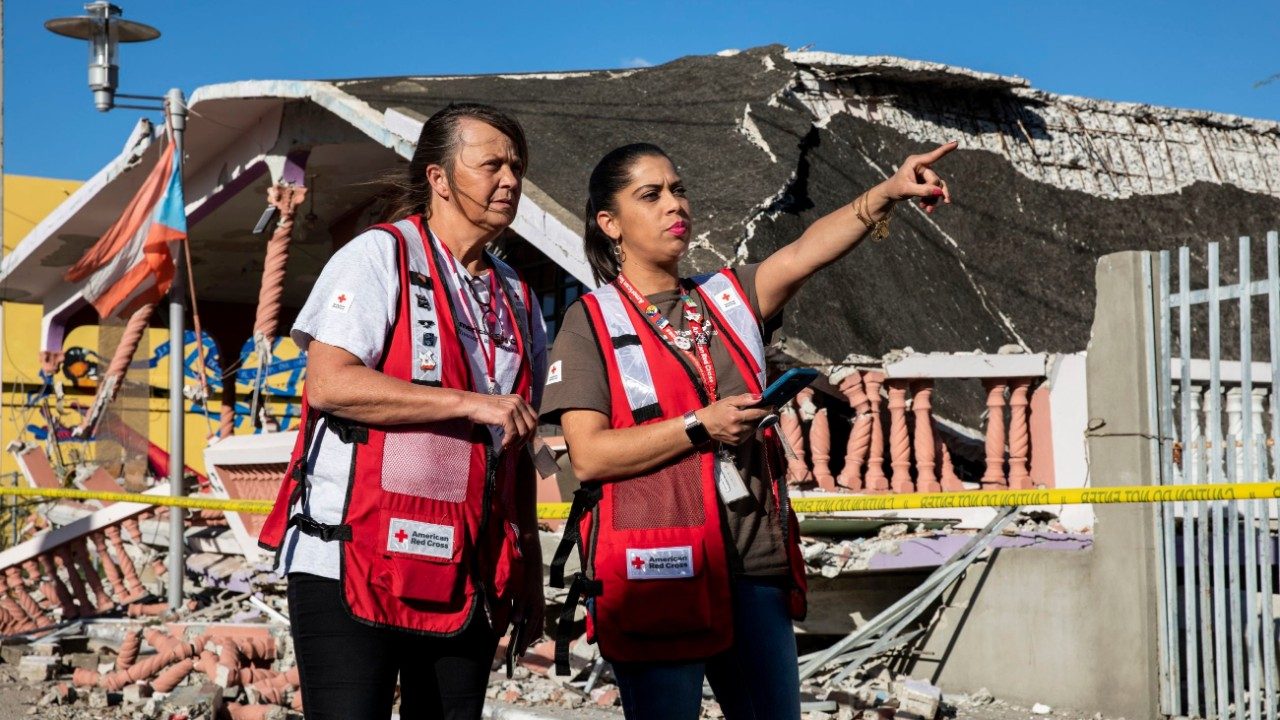 two Red Crossers in red vests stand outside a destroyed home